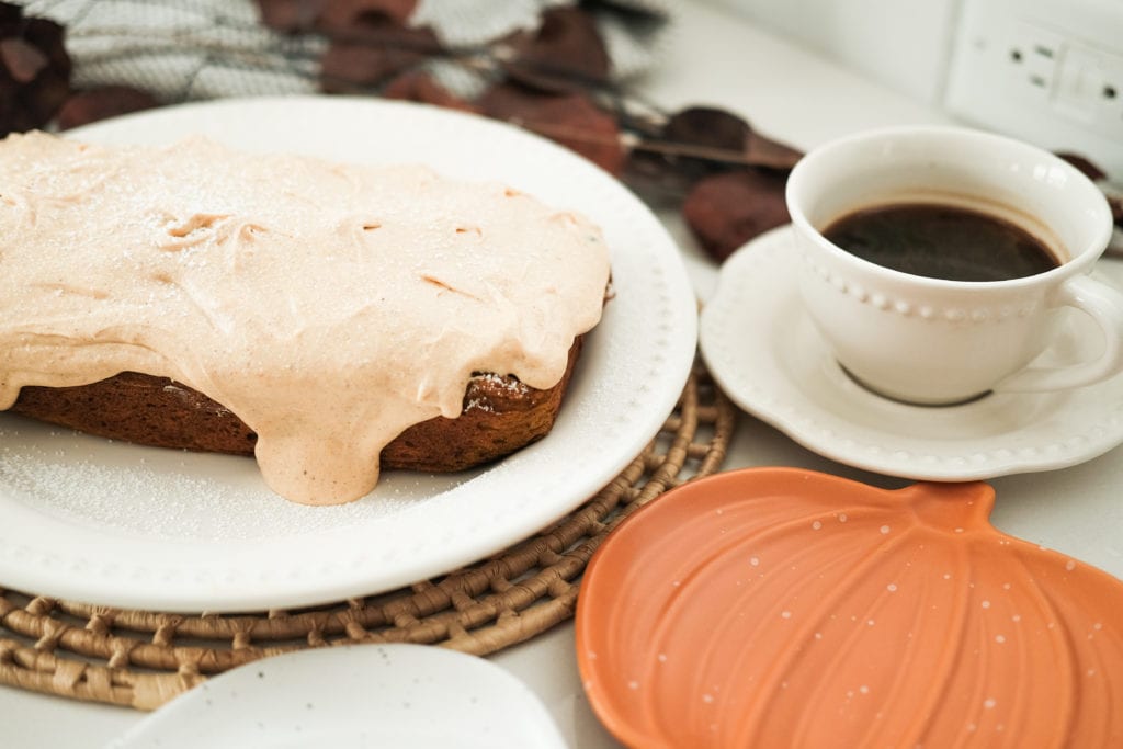 a frosted pumpkin carrot cake on a white plate, a pumpkin shaped plate next to it, and a coffee cup on a saucer filled with coffee 