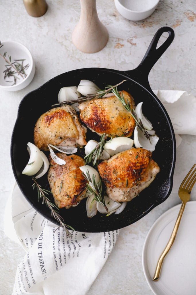 4 chicken thighs in cast iron skillet with linen and gold fork 
