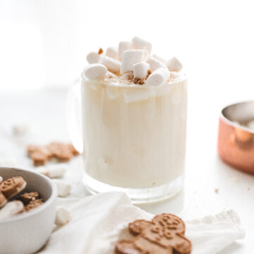 cup of white hot chocolate topped with marshmallows with gingerbread men garnish laying next to it.