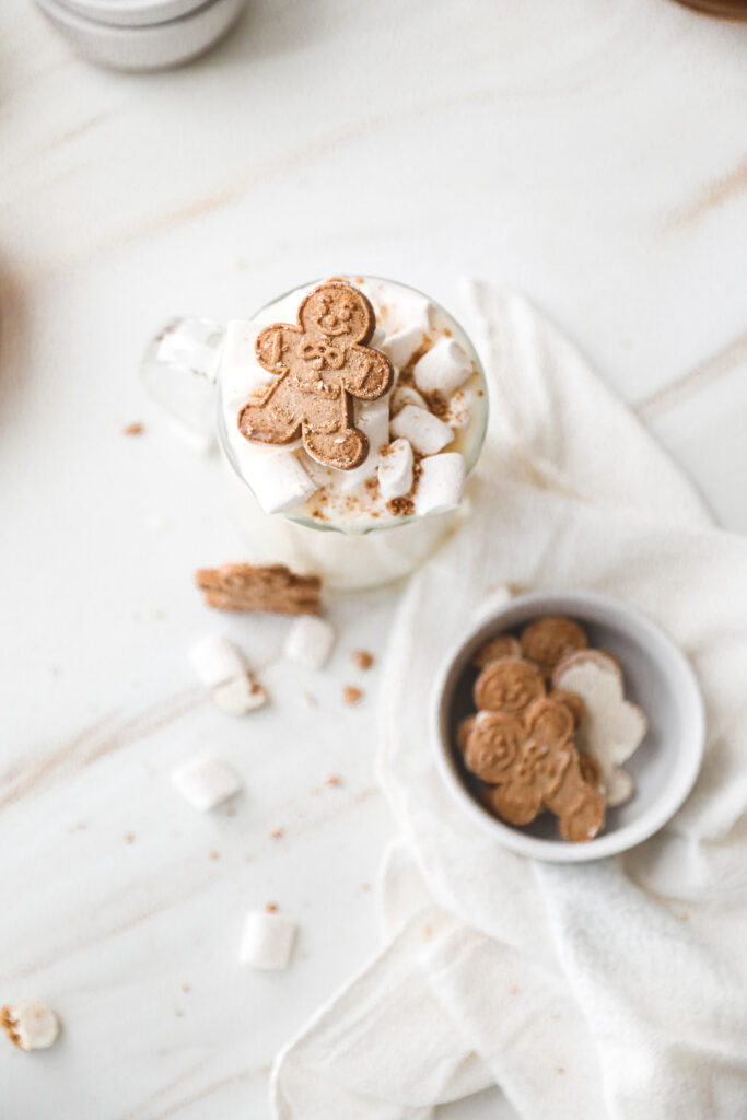 cup of white hot chocolate topped with marshmallows and gingerbread men with a white linen draped around and scattered ingredients