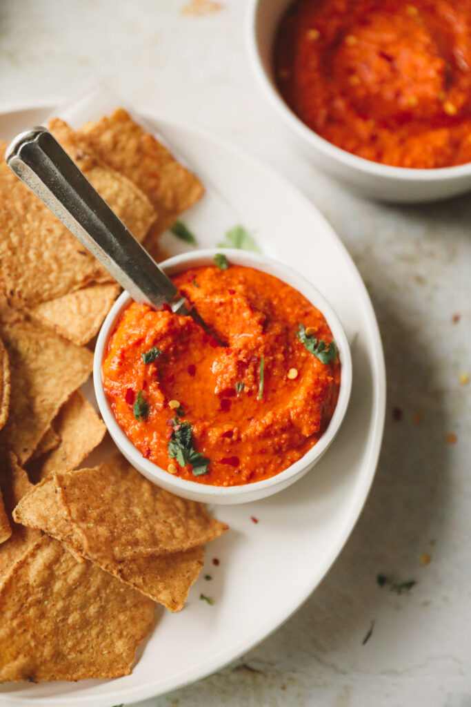 romesco dip in a white bowl with a spoon surrounded by corn chips on a white plate
