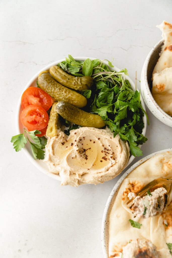 plate of hummus, parsley, pickles, and tomato on a white background 
