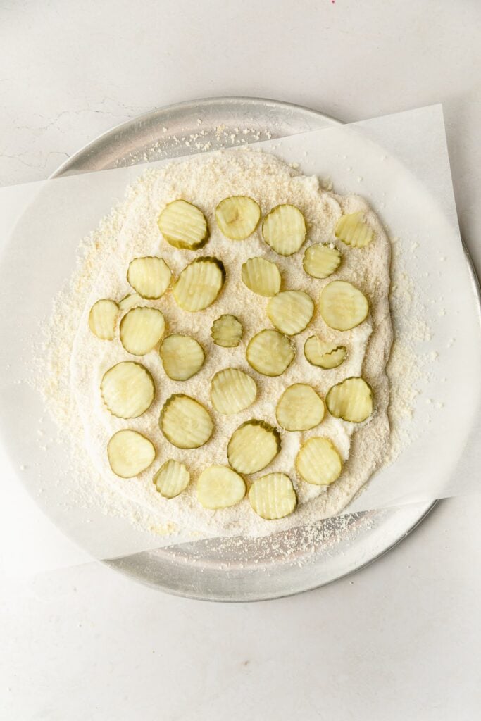 pizza dough rolled out into a circle topped with garlic sauce, parmesan cheese, and pickles