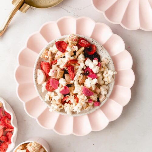 pink party Chex mix on a pink scalloped plate with other pink scalloped plates surrounding it