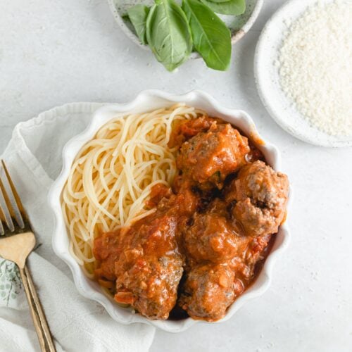 a scalloped edge plate with spaghetti, vodka sauce and meatballs. Surrounded by fresh basil and parmesan in white dishes