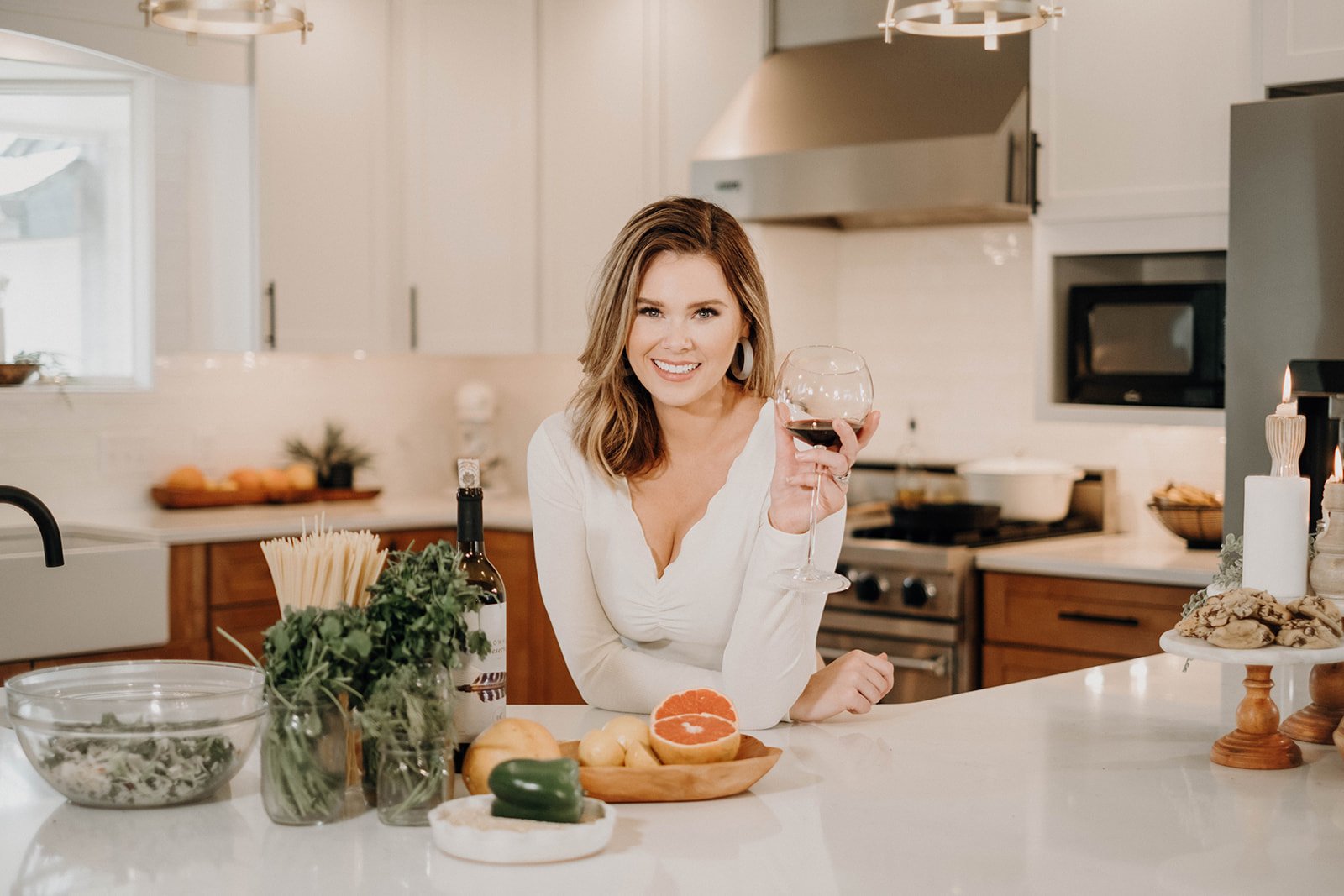 Sara ayes holding wine in a white kitchen with fresh ingredients around her