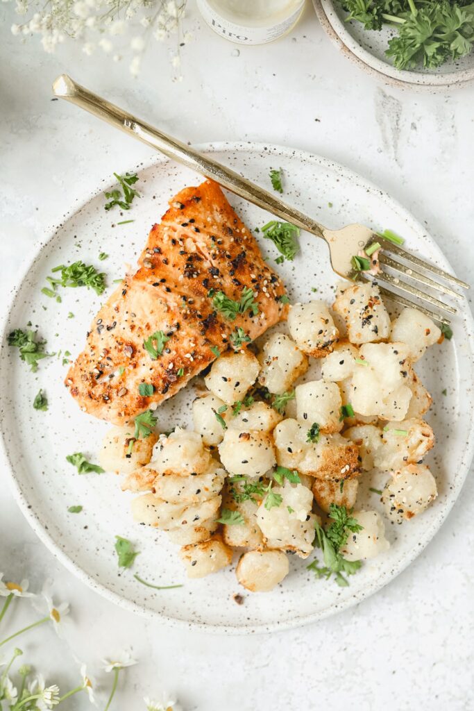 white plate with fried gnocchi and a salmon filet with a gold fork next to it.