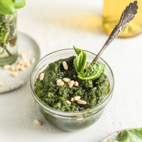 glass jar of pesto with spoon. Pesto is topped with pine nuts and basil leaves.