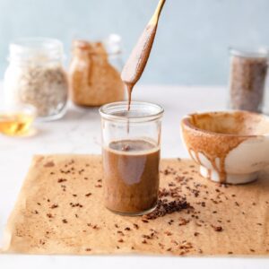 glass jar of granola butter on parchment paper with a knife drizzling granola butter into it