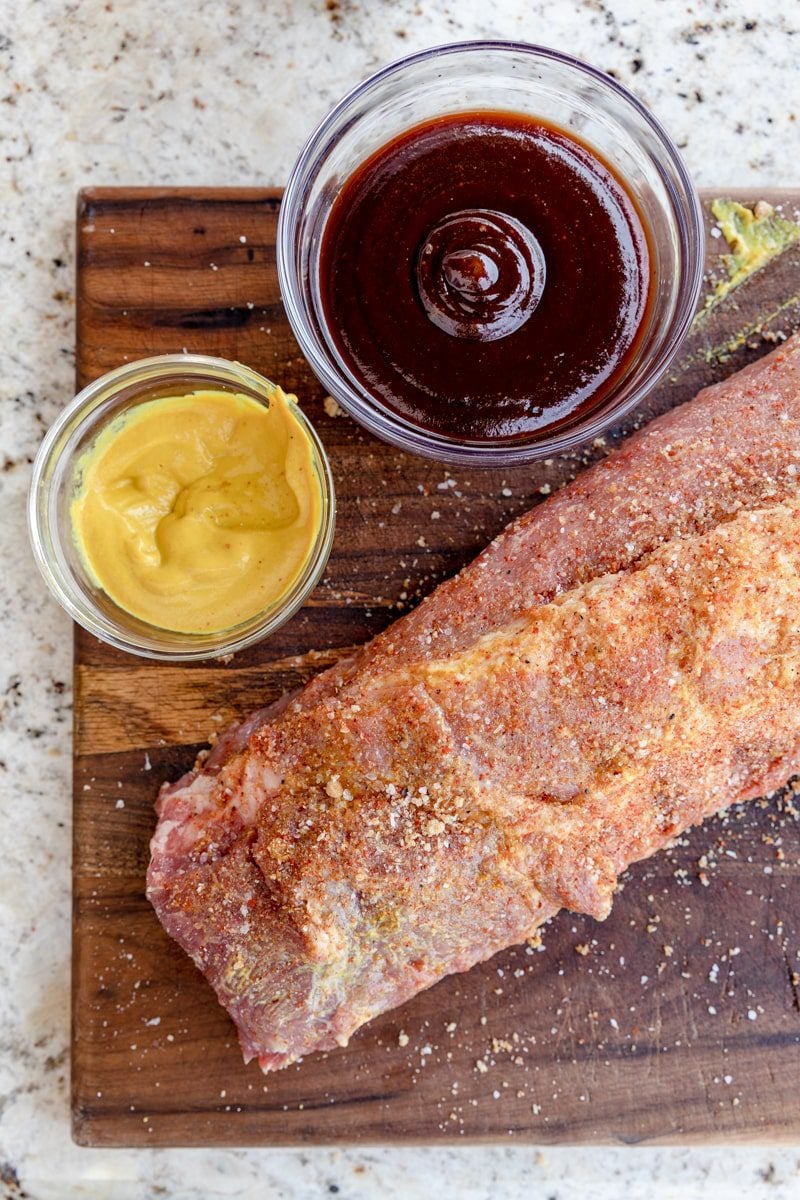 baby back ribs covered in yellow mustard and dry rub on a cutting board
