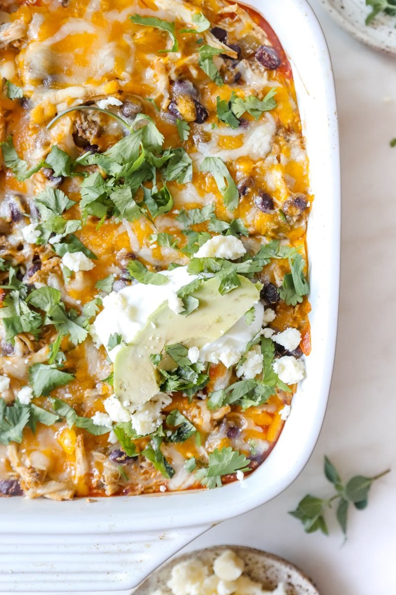 photo of the corner of a chicken enchilada casserole dish garnished with cilantro, sour cream, and avocado.