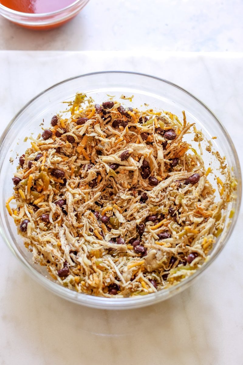 shredded rotisserie chicken, beans, seasoning, and cheese mixed together in a glass mixing bowl