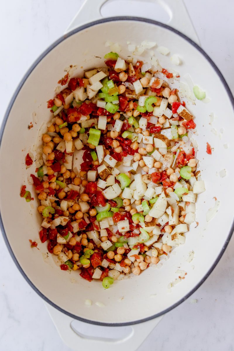 white dutch oven with tomato, potato, celery, onion, and spiced sautéed and stirred together