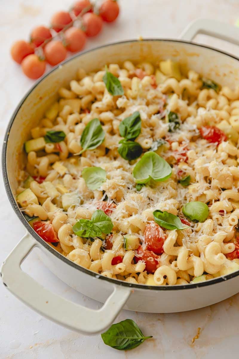 White skillet of creamy vegetable pasta garnished with basil.